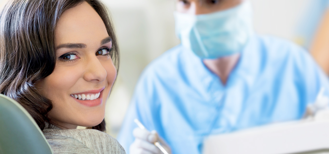Eine Frau auf dem Zahnarztstuhl sieht über die Schultern direkt in die Kamera und grinst.